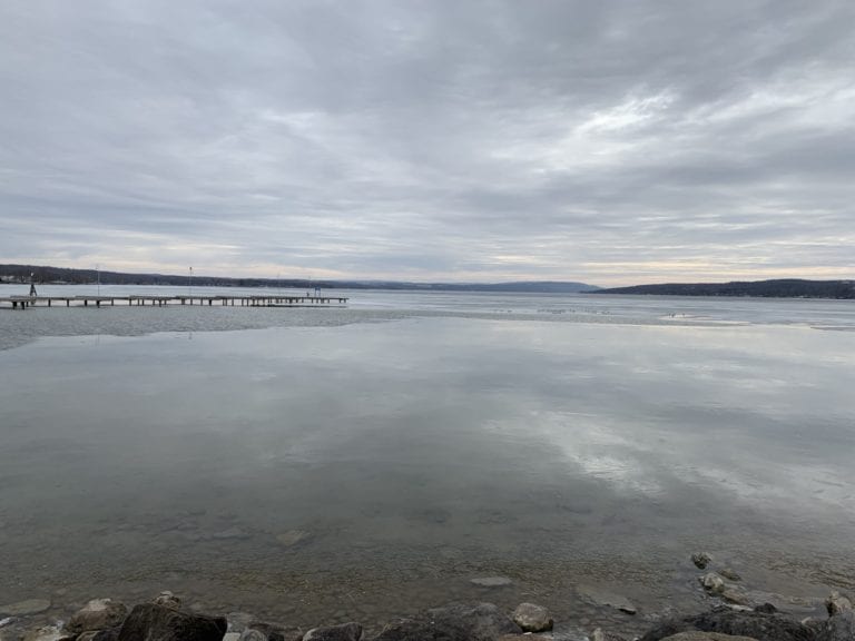 View of Canandaigua Lake over still, reflective waters with pier jutting out and hills in distance on a moody day in the Finger Lakes
