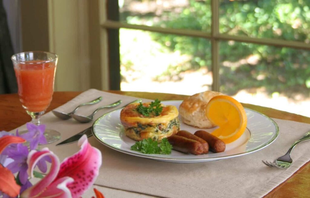 A plate with an egg muffin, two sausages, a biscuit, and an orange slice, next to a glass of juice.