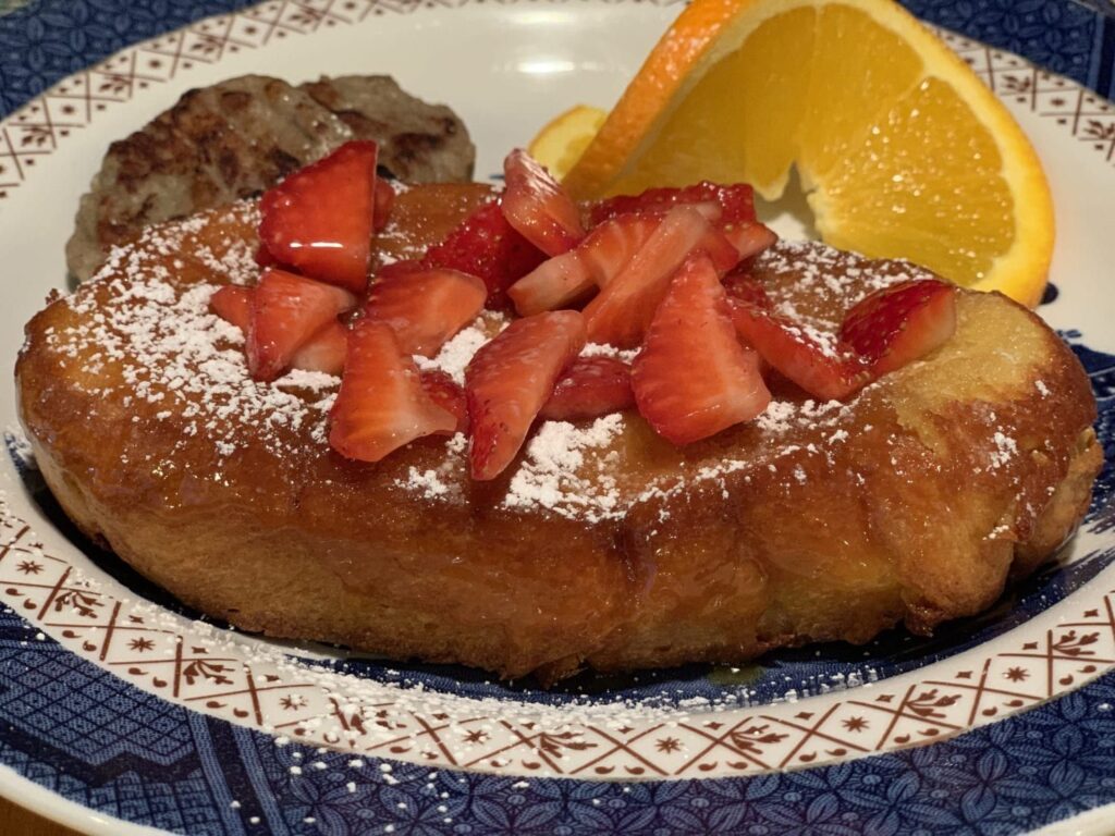 French toast topped with strawberries and powdered sugar, served with a sausage patty and orange slices.