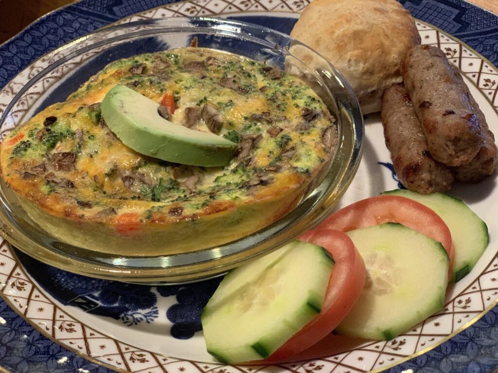 A vegetable frittata topped with avocado, served with two sausage links, a dinner roll, and cucumber and tomato slices.