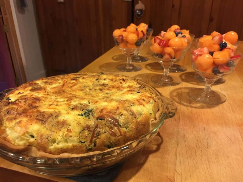 A quiche in a glass pie dish next to several glass cups filled with melon and berry fruit salad.