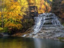 Buttermilk Falls gently flowing down steps of rock during fall foliage with yellow trees and still pond below