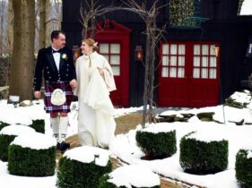 Bride in a white lace wedding gown and cape and groom in a traditional kilt looking at each other lovingly while walking from the Barn Suite to the Gathering Room at the 1795 Acorn Inn Bed and Breakfast on a winter's day with snow covered grounds