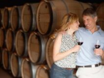 Couple dressed in summer clothing tasting red wine with wine barrels in the backgroud.